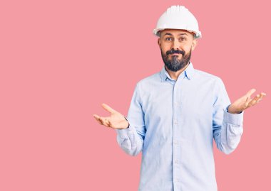 Mimar şapkası takan genç yakışıklı, kafası karışmış ve kolları ve elleri havada. şüphe kavramı. 