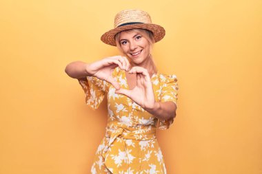 Sarışın, güzel bir kadın tatilde. Sarı arka planda şapkası ve elbisesi var. Aşkla gülümsüyor, elleriyle kalp sembolü yapıyor. Romantik konsept.