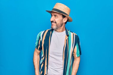 Orta yaşlı yakışıklı turist gömlekli ve şapkalı, mavi arka planda, rahat bir görünüm ve kendine güvenen bir gülümseme takınıyor..