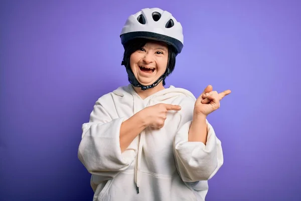 Young down syndrome cyclist woman wearing security bike helmet over purple background smiling and looking at the camera pointing with two hands and fingers to the side.