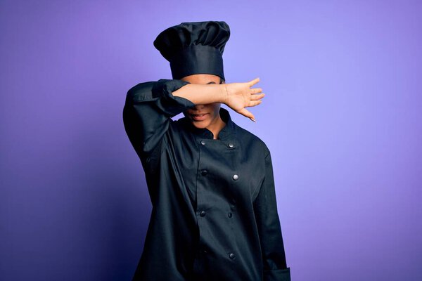 Young african american chef girl wearing cooker uniform and hat over purple background covering eyes with arm, looking serious and sad. Sightless, hiding and rejection concept