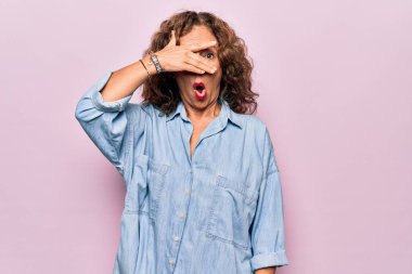 Orta yaşlı güzel bir kadın pembe arka planda sıradan bir kot gömlek giyer. Şoka girer, yüzünü ve gözlerini kapar ve utanmış bir ifadeyle parmaklarından bakar..