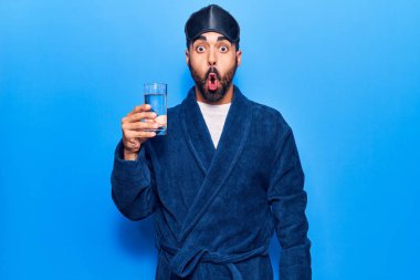 Uyku maskesi takan ve bornoz giyen genç İspanyol adam su içerken korkmuş ve şaşırmış bir şekilde ağzı açık kalmış, inançsız bir yüz. 