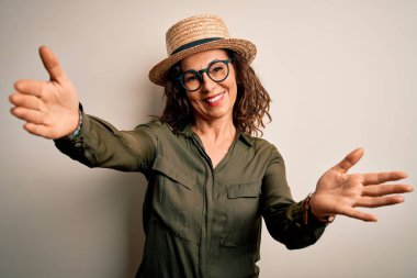 Orta yaşlı esmer kadın, gözlüklü ve şapkalı, izole edilmiş beyaz arka planda duruyor. Kameraya bakıyor ve sarılmak için kollarını açmış gülümsüyor. Mutluluğu kucaklayan neşeli bir ifade..