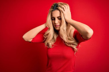 Genç ve güzel sarışın kadın, günlük tişört giyip izole edilmiş kırmızı arka planda dikiliyor. Ağrı ve migrenden dolayı baş ağrısı ve stresten muzdarip. Ellerini başının üstüne koy..