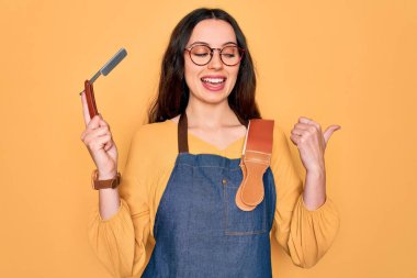 Önlük takan genç, güzel bir berber kadın sarı arka planda jilet tutuyor ve gülümseyen bir gülümsemeyle baş parmağıyla onu işaret ediyor.