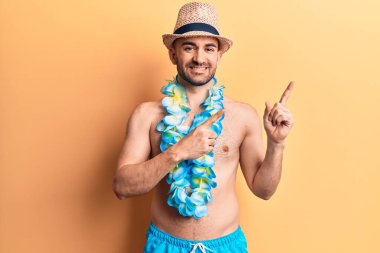 Yakışıklı, kel, mayo giymeyen, Hawaii zambağı takan, gülümseyen ve kameraya bakıp iki elini ve parmağını yana doğru gösteren bir adam.. 