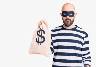 Young handsome man wearing burglar mask holding money bag with dollar symbol thinking attitude and sober expression looking self confident  clipart