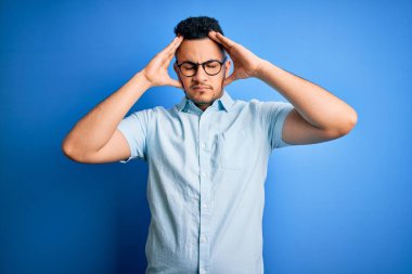 Yaz gömleği ve gözlük takan genç yakışıklı bir adam izole edilmiş mavi arka planda ağrı ve migrenden dolayı çaresiz ve stresli bir baş ağrısından muzdarip. Ellerini başının üstüne koy..