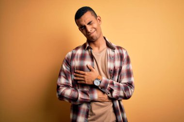 Genç, yakışıklı, Afro-Amerikan bir adam günlük gömleğini giymiş sarı arka planda, midesi üstünde duruyor çünkü hazımsızlık, acı verici bir hastalık, kendini iyi hissetmiyor. Ache kavramı.