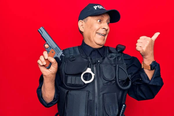 Senior man with grey hair wearing police uniform holding gun pointing thumb up to the side smiling happy with open mouth