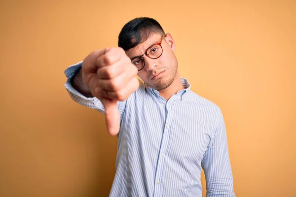 黄色の背景にオタク眼鏡をかけた若いハンサムなヒスパニック系のビジネスマンは 親指を下にジェスチャーで拒絶と否定を示す不満と怒りを見ています 悪い表現 — ストック写真