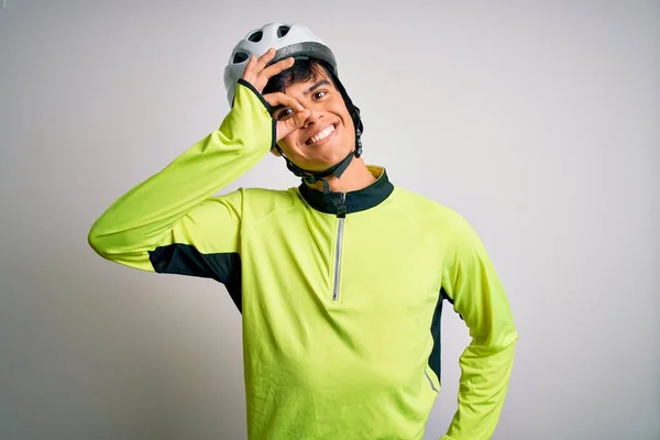 Young handsome cyclist man wearing security bike helmet over isolated white background doing ok gesture with hand smiling, eye looking through fingers with happy face.