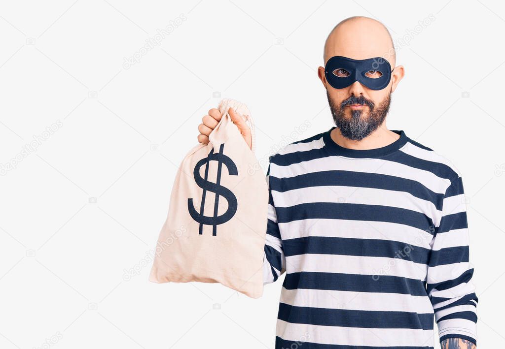 Young handsome man wearing burglar mask holding money bag with dollar symbol thinking attitude and sober expression looking self confident 
