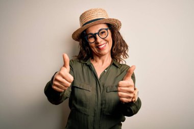Orta yaşlı esmer kadın gözlüklü ve şapkalı, izole edilmiş beyaz arka planda duruyor. Elle olumlu jest yapıyor, gülümsüyor ve başarı için mutlu. Kazanan jest.