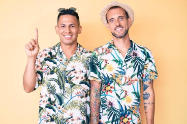 Yaz şapkası ve Hawaii tişörtü giyen iki genç gay bir numarayı gösterip gösterirken kendinden emin ve mutlu bir şekilde gülümsüyor.. 