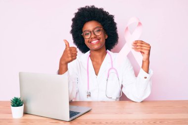 Genç Afro-Amerikan kadın doktor üniforması giyiyor pembe kanser kurdelesi taşıyor mutlu ve pozitif gülümsüyor, başparmağıyla mükemmel ve onay işareti yapıyor. 