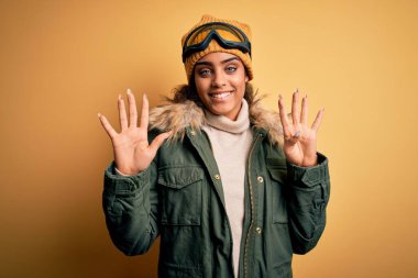 Genç Afrikalı Afro-Amerikan kayakçı kız kar sporu giysisi ve kayak gözlüğü takıyor 9 numarayı gösterip gösterirken kendine güvenen ve mutlu gülümseyen..