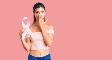 Genç ve güzel bir kadın elinde kanser farkındalığı olan pembe kurdeleli ağzı elle kaplayan, şok olmuş ve hata yapmaktan korkan bir kadın. şaşırmış ifade 