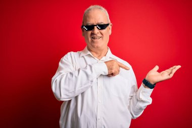 İzole edilmiş kırmızı arka planda komik güneş gözlüğü takan orta yaşlı beyaz adam kameraya hayret ediyor ve gülümsüyor..