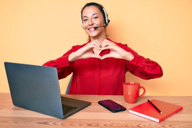 Çağrı merkezindeki operatör kulaklığı takan genç esmer kadın kalp sembolü ve şekillerini elleriyle gösteren aşkla gülümsüyor. romantik kavram. 