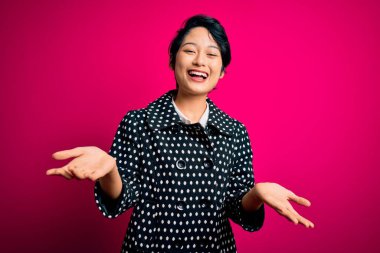 Genç, güzel Asyalı bir kız, günlük ceket giyiyor pembe arka planda dikiliyor, gülümsüyor, yardım eli uzatıyor ve kabul ediyor..