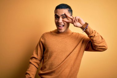 İzole edilmiş sarı arka plan üzerinde dikilen Brezilyalı genç adam barış sembolü yapıyor. Yüzünde parmaklarıyla gülümsüyor ve zaferini gülümsüyor.