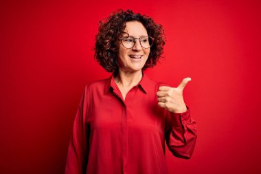 Orta yaşlı, kıvırcık saçlı, rahat gömlekli, kırmızı arka planda gözlüklü, mutlu yüzlü ve baş parmağıyla yüzünü gösteren güzel bir kadın..