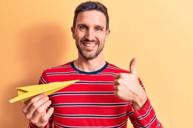 Sarı kağıt uçak tutan yakışıklı genç adam izole edilmiş arka planda duruyor ve gülümsüyor mutlu ve pozitif, başparmak yukarı, mükemmel ve onay işareti yapıyor.