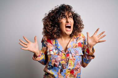Genç, kıvırcık Arap kadın çiçek rengi gömlek giyiyor beyaz arka planda duruyor deli deli deli bağırıyor ve agresif bir ifadeyle bağırıyor ve kollarını kaldırıyor. Hayal kırıklığı konsepti.