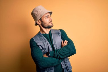 Tatilde sakallı yakışıklı turist sarı arka planda kaşif şapkası takıyor kolları çapraz ve kendinden emin bir şekilde yana bakıyor.