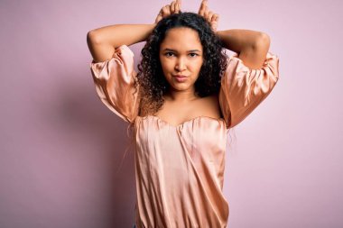 Kıvırcık saçlı, pembe tişörtlü genç bir kadın pembe arka planda dikilmiş, boynuzlarını başlarının üzerinde gezdirirken komik hareketler yapıyor.