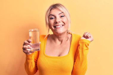 Sarışın, güzel bir kadın, izole edilmiş sarı arka planda bir bardak su içiyor gururla bağırıyor, zaferi kutluyor ve kollarını kaldırarak başarıyı kutluyor.