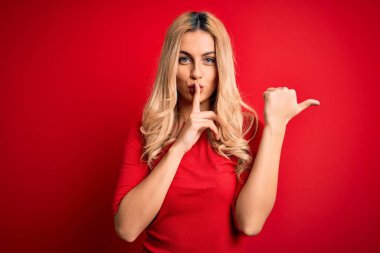 Genç ve güzel sarışın bir kadın, günlük tişört giyip kırmızı arka planda dikiliyor ve parmakları dudaklarını yana doğru gösteren parmaklarıyla sessiz olmak istiyor. Sessizlik ve gizli konsept.