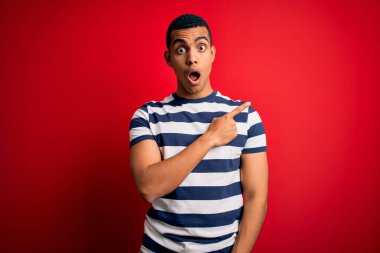 Yakışıklı Afro-Amerikan erkeği kırmızı arka planda dikilen sıradan çizgili tişört giyiyor. Parmakla işaret ediyor, ağzı açık hayret içinde..
