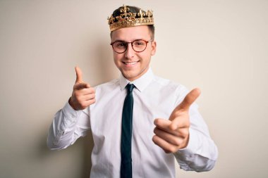 İzole edilmiş arka planda altın tacını takmış yakışıklı beyaz iş adamı mutlu ve komik yüzlü parmaklarını kameraya doğrultuyor. İyi enerji ve titreşimler.