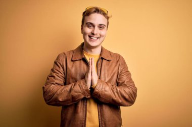 Genç, kızıl saçlı, günlük deri ceket giyen, izole edilmiş sarı arka planda dua eden ellerini birleştirip af dileyen, kendinden emin gülümseyen bir adam..