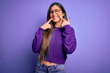 Genç, güzel, zeki bir kadın mor bir arka planda gözlük takıyor açık ağızla gülümsüyor, parmakları işaret ediyor ve zorla neşeli bir gülümseme sergiliyor.