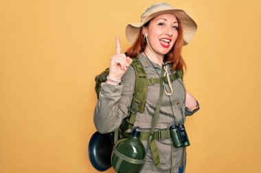 Sırt çantalı, sarı arka planda şapka takan ve başarılı bir fikri işaret eden kızıl saçlı genç bir kadın. Heyecanlı ve mutlu. Bir numara..