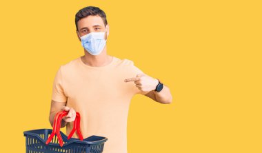 Alışveriş sepeti ve sağlık maskesi takmış yakışıklı genç adam gülümseyen mutlu ve gururlu birini işaret ediyor. 