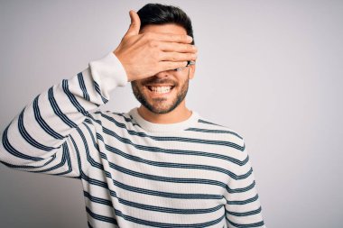 İzole edilmiş beyaz arka planda optometri gözlüğü takan sakallı yakışıklı genç bir adam. Gülümsüyor ve elleri yüzünde şaşkınlıktan gözleri kapatıyordu. Kör kavram.