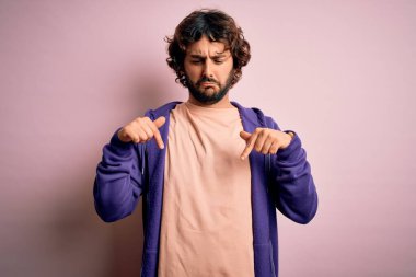 Genç, yakışıklı, sakallı, pembe arka planda sıradan bir kazak giyen üzgün ve üzgün görünen, parmaklarıyla yön gösteren mutsuz ve depresif bir adam..