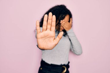 Genç Afro-Amerikan kadın balıkçı yaka kazak ve pembe arka plan üzerine gözlük takıyor gözleri ellerle örtüyor ve üzgün ve korku ifadeleriyle jestleri durduruyor. Utanç verici ve olumsuz bir kavram..