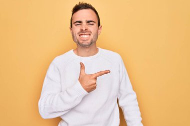 Mavi gözlü, sıradan bir süveter giyen, sarı arka planda duran, yüzünde mutlu ve doğal bir ifadeyle elini ve parmağını gösteren bir gülümseme olan genç yakışıklı adam.