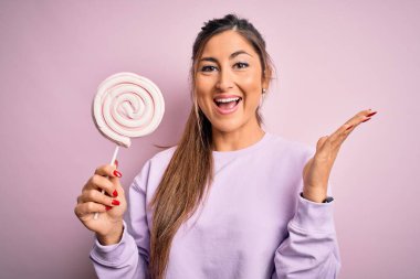 Genç, güzel esmer kadın pembe arka planda tatlı şekerler yiyor. Çok mutlu ve heyecanlı. Kazanan yüz ifadesi zaferi kutluyor.