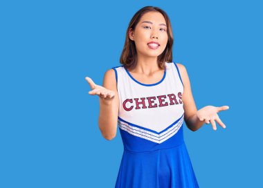 Genç, güzel Çinli kız ponpon kız üniforması giyiyor. Kollarını açıp gülümsüyor. Hoş geldin, olumlu ve kendinden emin selamlar. 