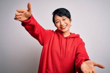 Beyaz arka planda kapüşonlu sıradan bir sweatshirt giyen genç, güzel Asyalı kız sarılmak için kollarını açmış kameraya bakıyor. Mutluluğu kucaklayan neşeli bir ifade..