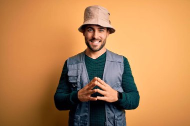 Tatilde sakallı yakışıklı turist sarı arka planda kaşif şapkası takıyor eller yan yana, gülümseyerek rahat ve neşeli. Başarı ve iyimser