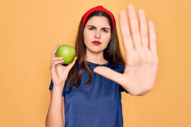 Genç ve güzel bir kız sarı arka planda taze organik yeşil elma yiyor. Açık elle dur işareti yapıyor. Ciddi ve kendinden emin bir ifadeyle.