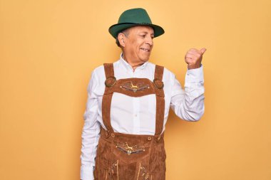 Sarı arka planda Alman geleneksel ahşap şenliği kıyafeti giyen yaşlı gri saçlı adam gülümsüyor ve başparmağıyla yan tarafa bakıyor..